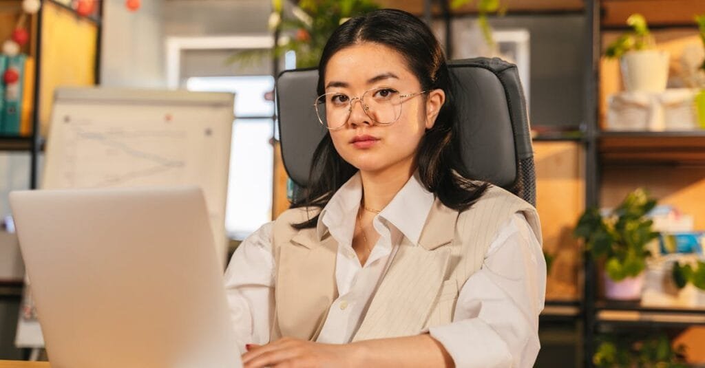 Confident businesswoman with glasses focusing on work using a laptop in a modern office setting.
