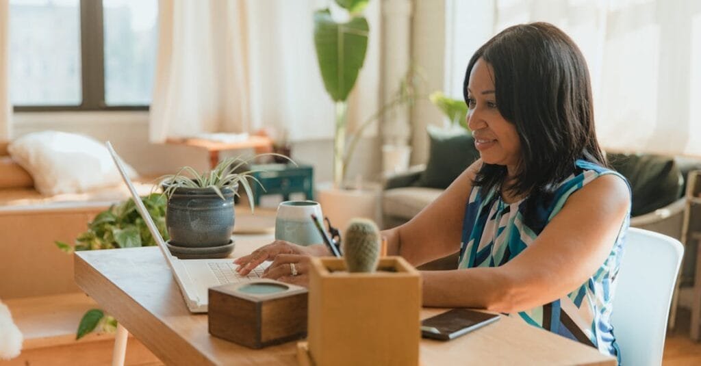 Woman working remotely at home, using laptop, surrounded by plants, creating a cozy workspace.
