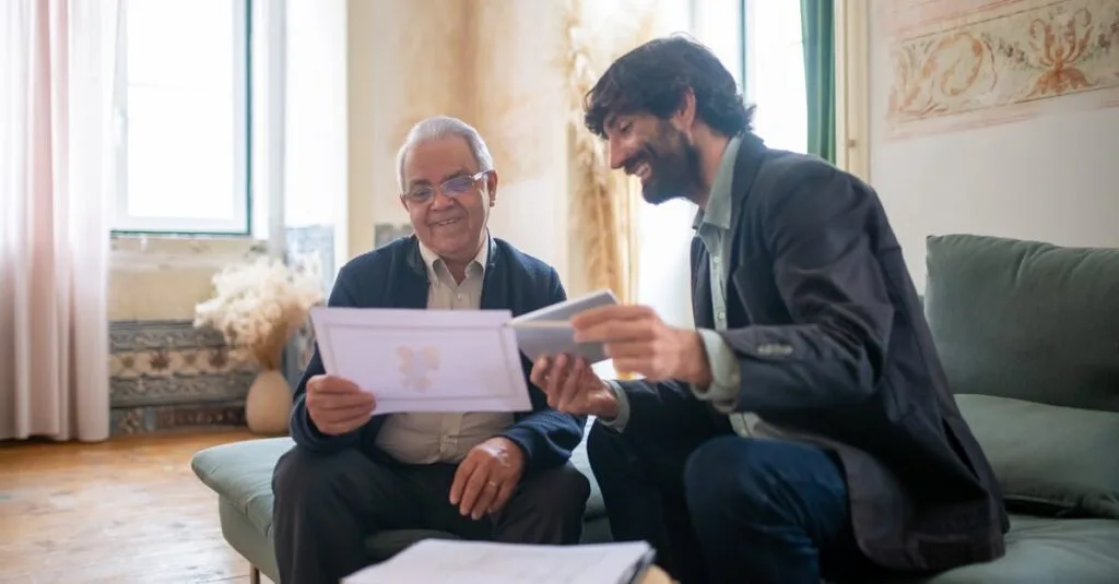 An elderly man and a younger man in a casual office setting, discussing documents with smiles.