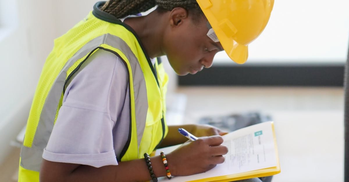 Focused female engineer in a safety helmet and vest writing on a clipboard indoors.