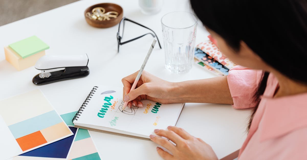 Close-up of a woman drawing and lettering in a notebook, showcasing creative handwriting and art supplies.