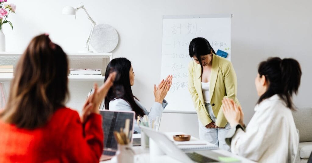Asian women in an office setting clapping for a colleague after a presentation with a whiteboard in the background.
