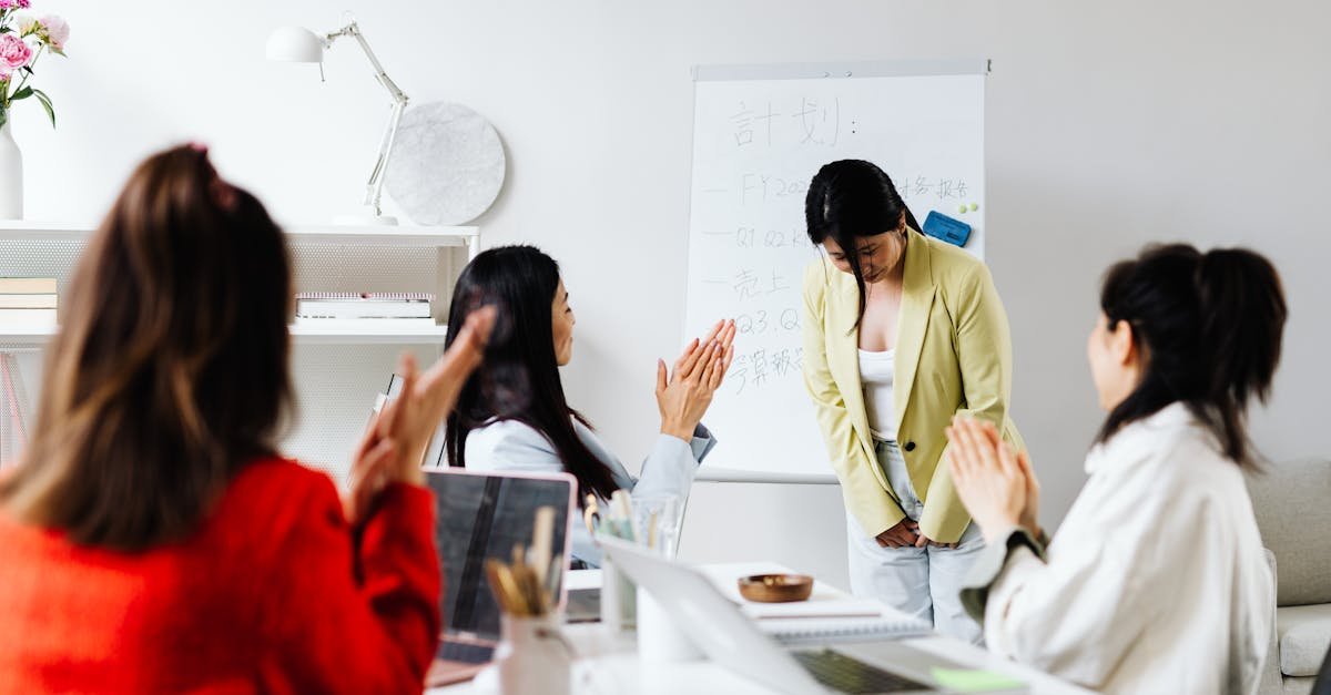 Asian women in an office setting clapping for a colleague after a presentation with a whiteboard in the background.