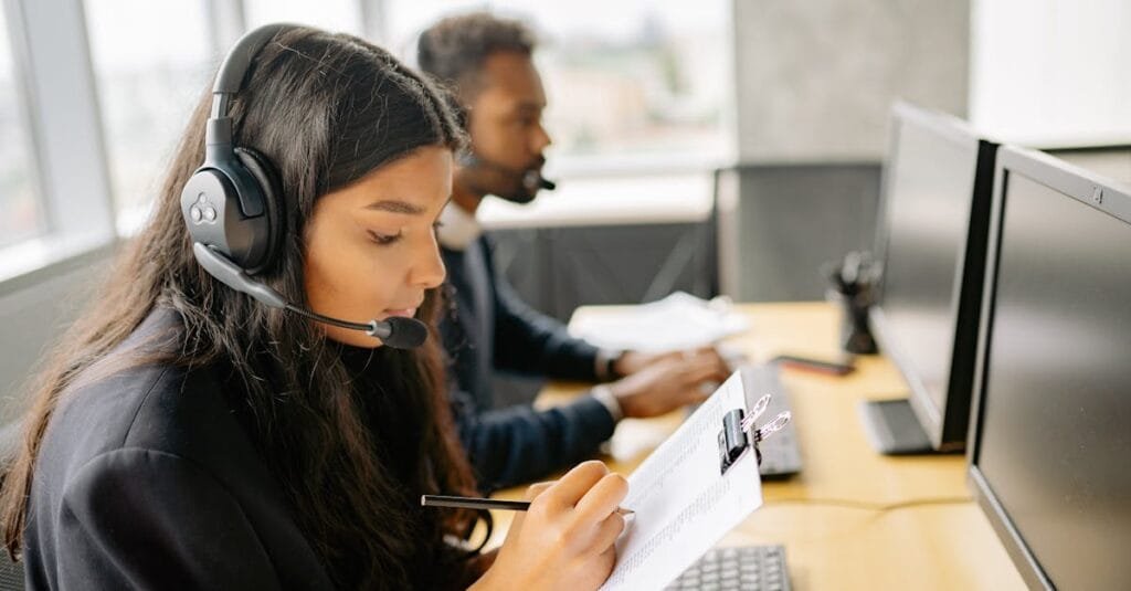 Two call center agents wearing headsets working at desks with computers in a modern office.