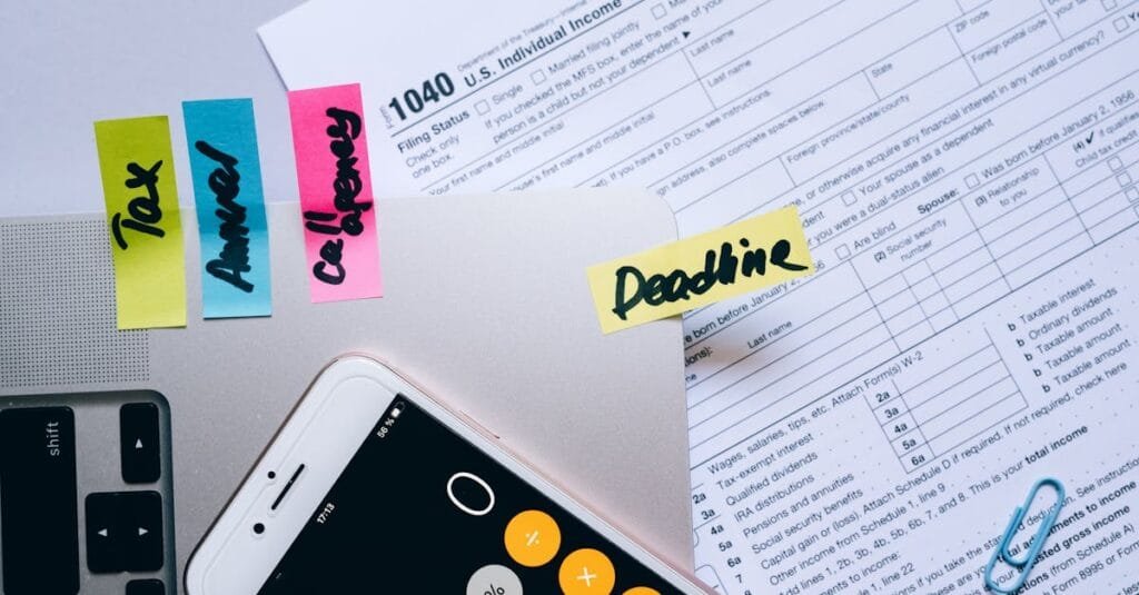 Office desk flat lay showing tax documents, calculator app on smartphone, sticky notes, and paperclips.