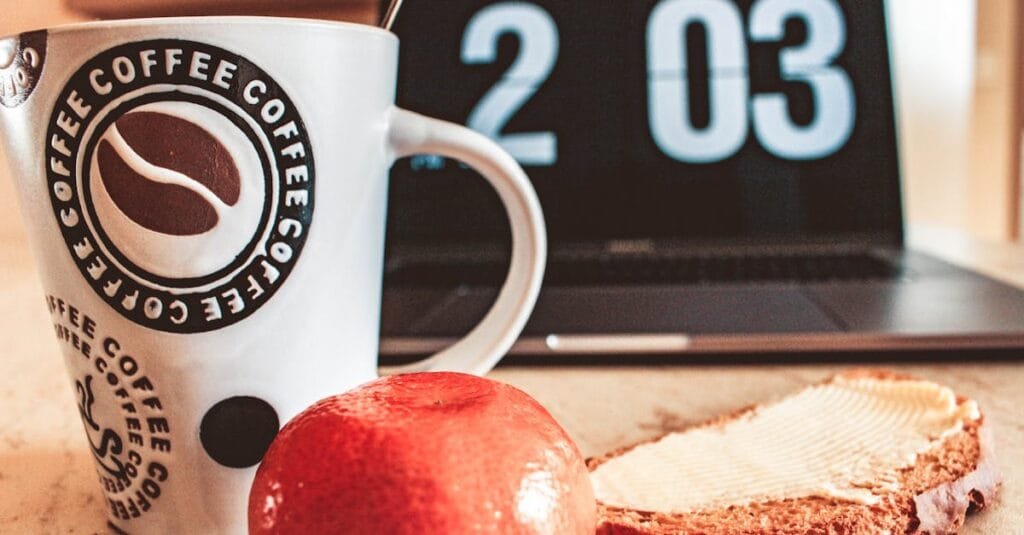 A cozy breakfast setup with coffee, bread, and fruit next to a laptop.