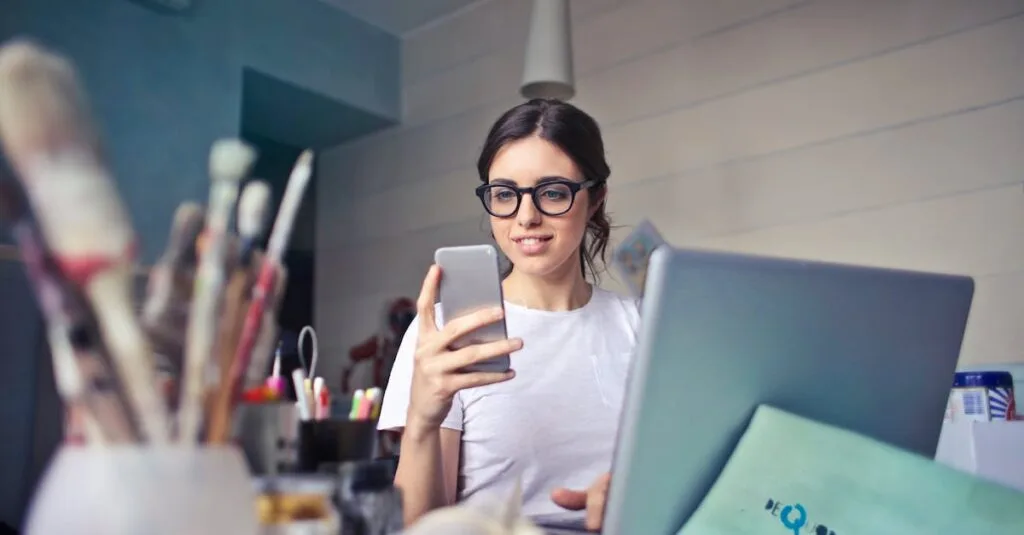 A woman using her phone at a desk, surrounded by art supplies and a laptop, in a creative workspace.