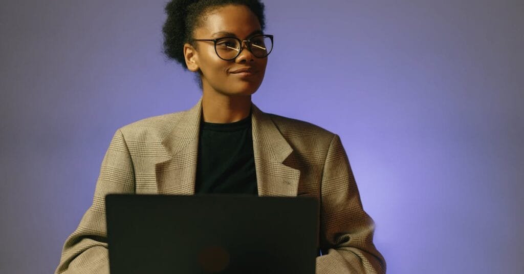 Portrait of a confident woman in a blazer working on a laptop, looking away thoughtfully in a studio setting.