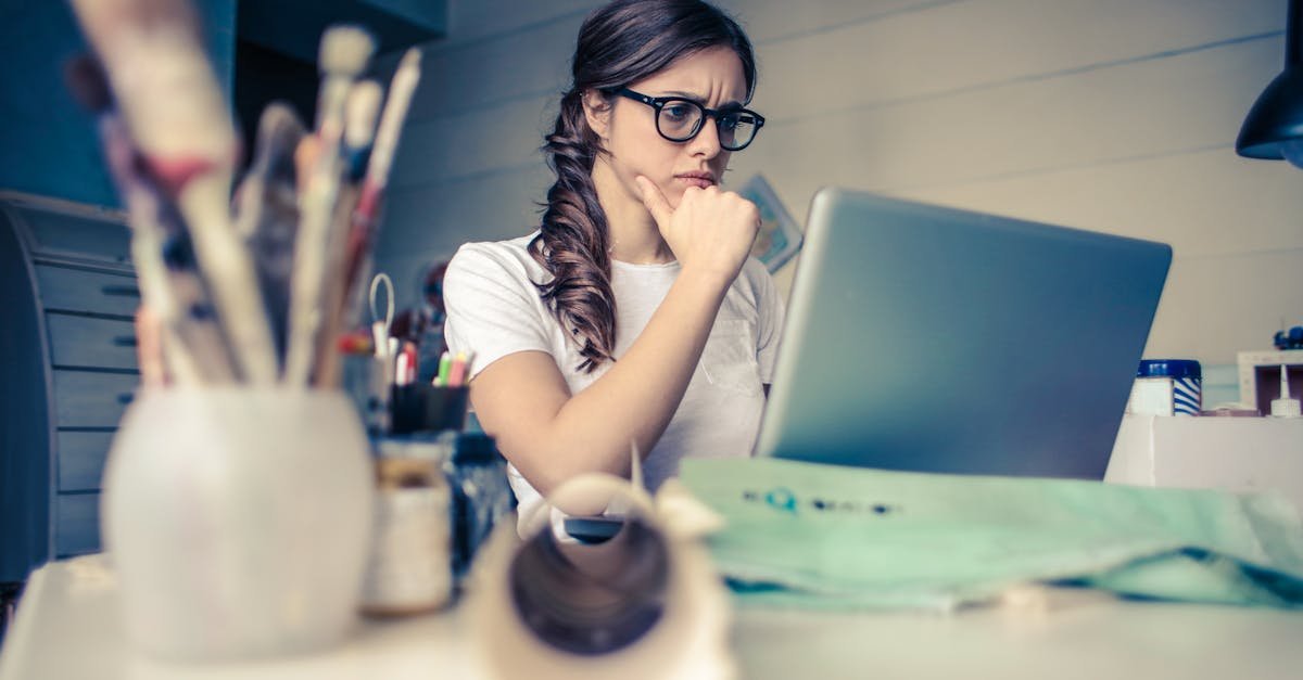 Young woman with glasses deeply focused on a laptop surrounded by art supplies in a home office.