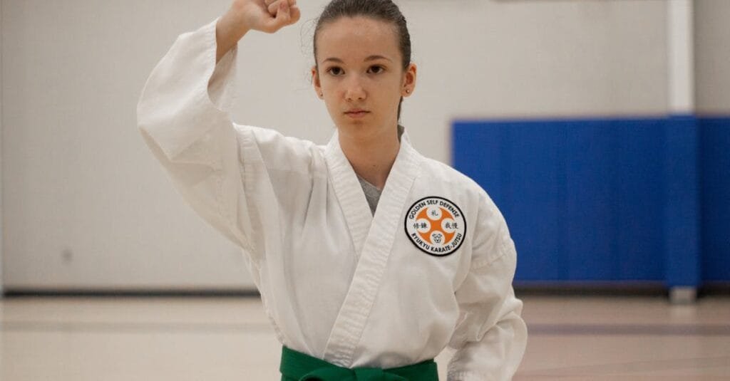 A young girl in a martial arts dojo practicing karate, wearing a green belt and a karategi.