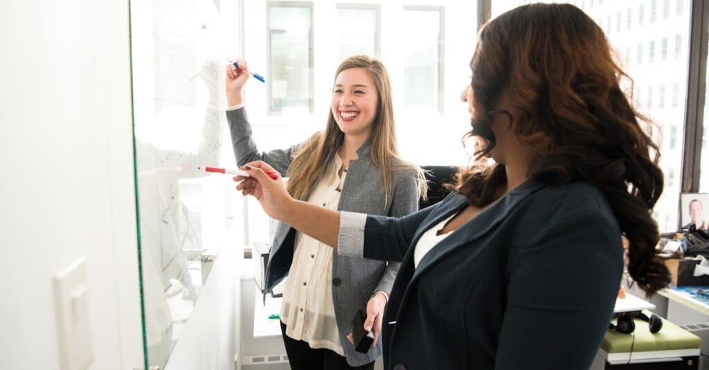 Two professional women discussing ideas on a whiteboard in a modern office setting.