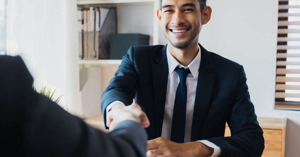 Confident businessman in suit shaking hands at office desk, symbolizing successful partnership.