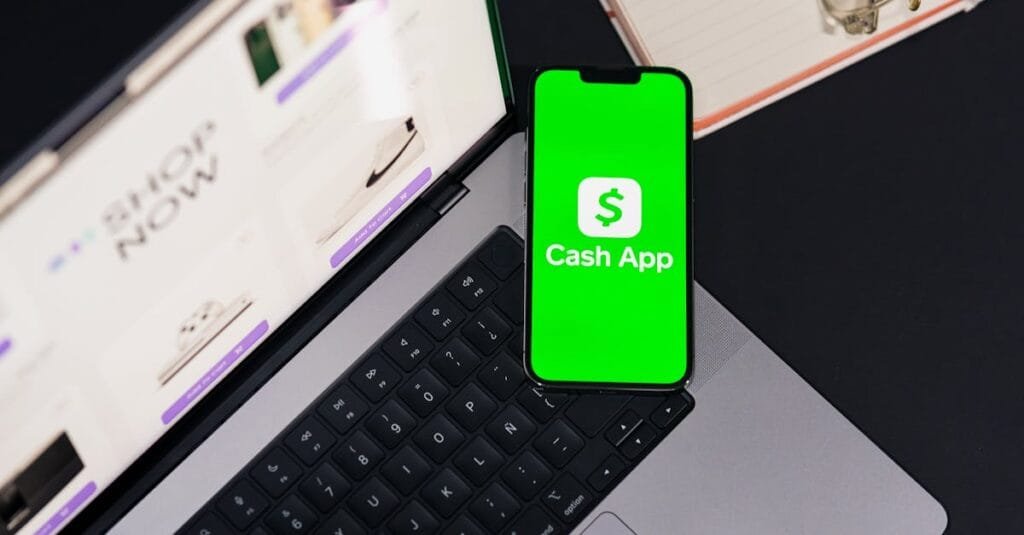 Smartphone showing Cash App screen on laptop keyboard, next to glasses and notebook.