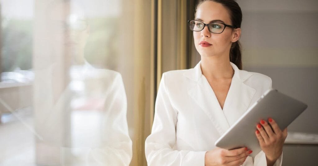 Businesswoman in formal attire thinking beside a window with a tablet.