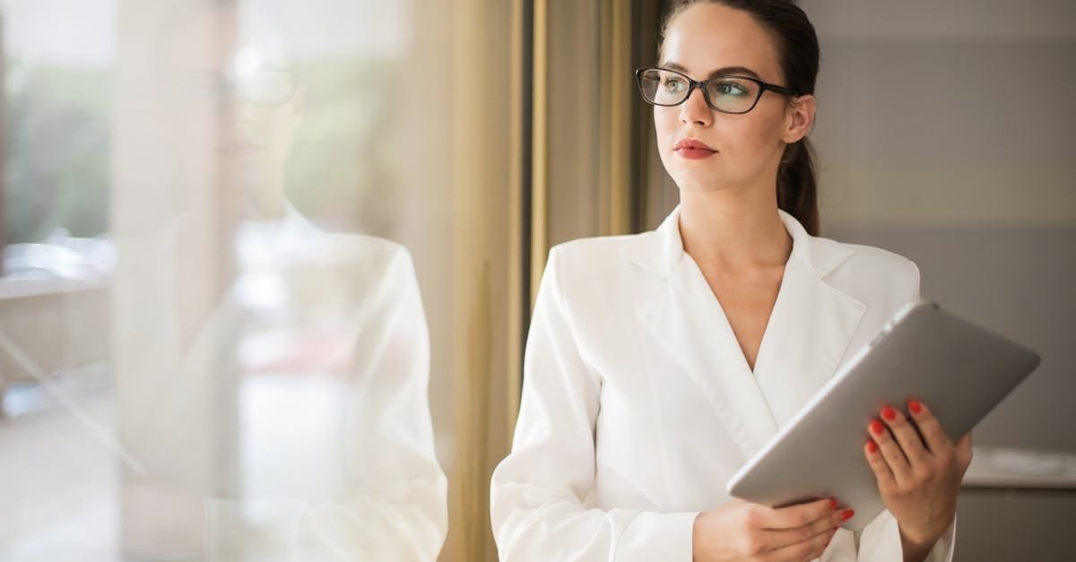 Businesswoman in formal attire thinking beside a window with a tablet.