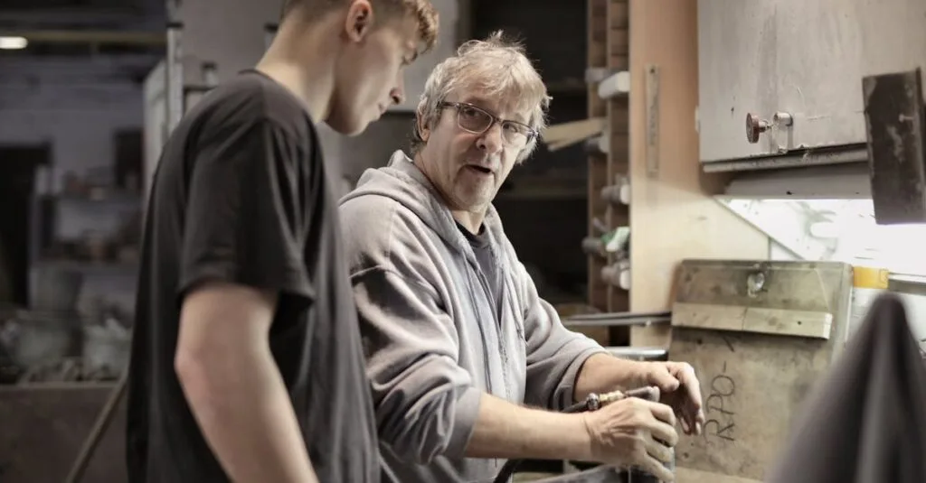 Side view of senior foreman in eyeglasses showing to trainee how handling detail in workshop
