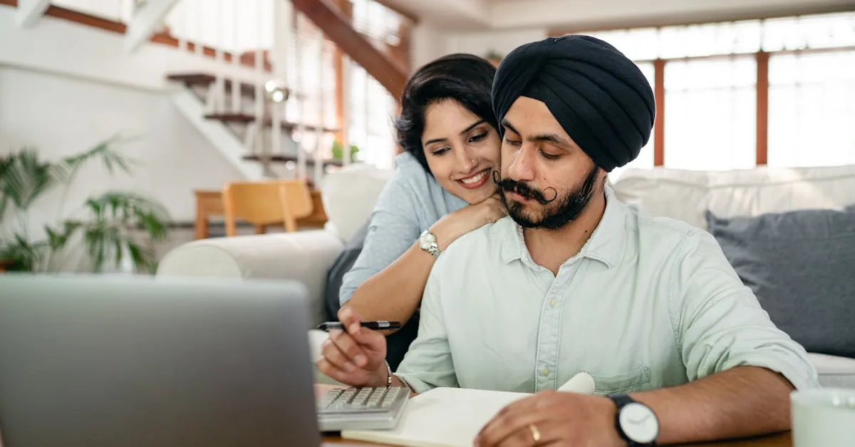 Cheerful young Indian woman cuddling and supporting serious husband working at home with laptop and counting on calculator