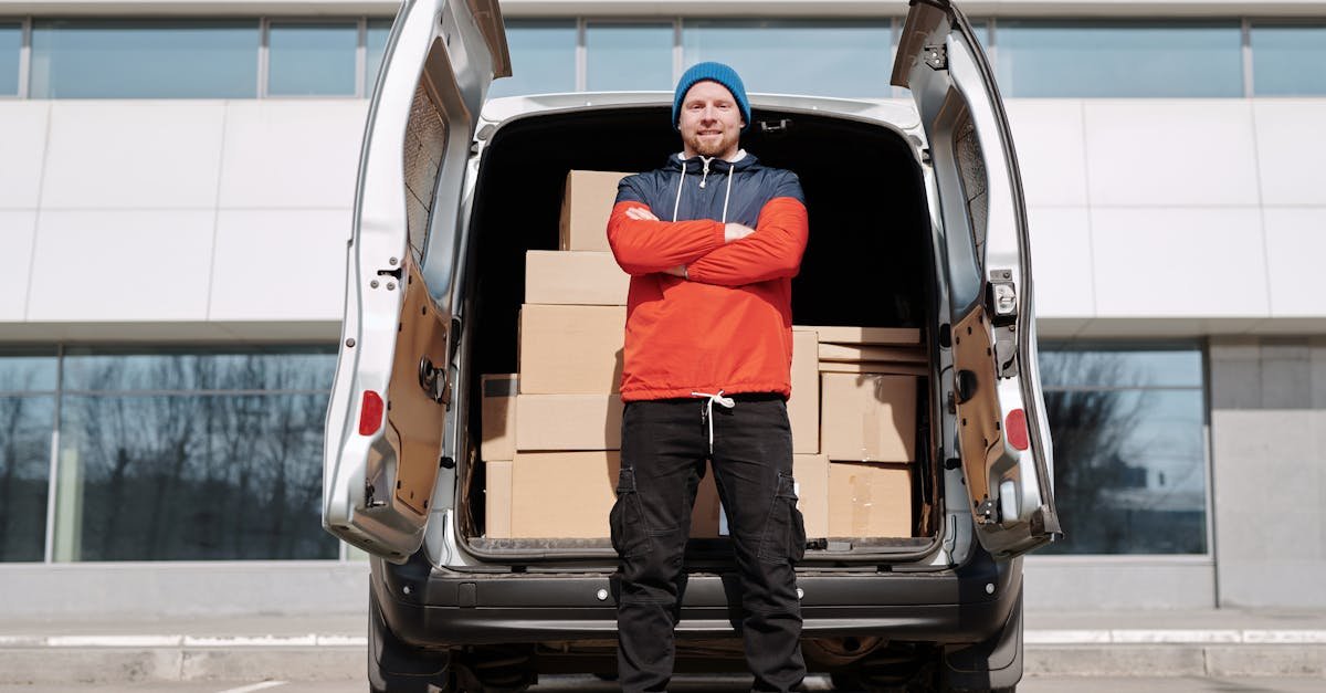 Man in red jacket and blue beanie stands by van of boxes in the city, ready for delivery.