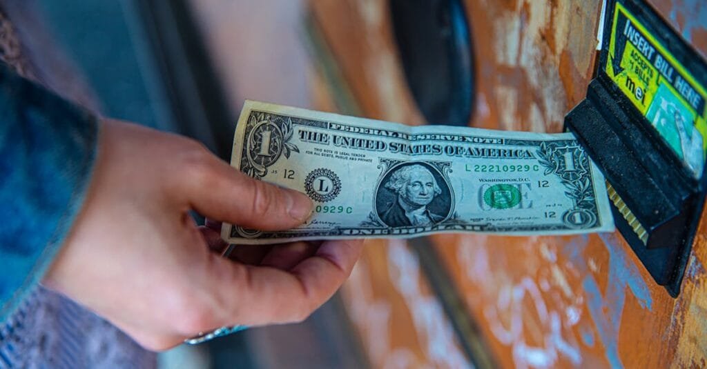 A person inserting a US dollar bill into a vending machine slot, capturing a financial transaction moment.