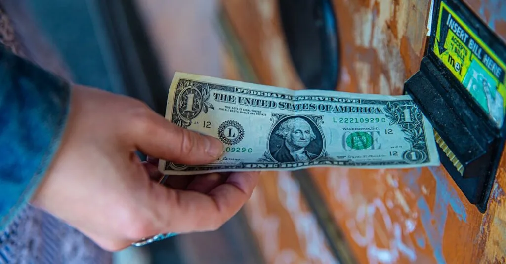 A person inserting a US dollar bill into a vending machine slot, capturing a financial transaction moment.