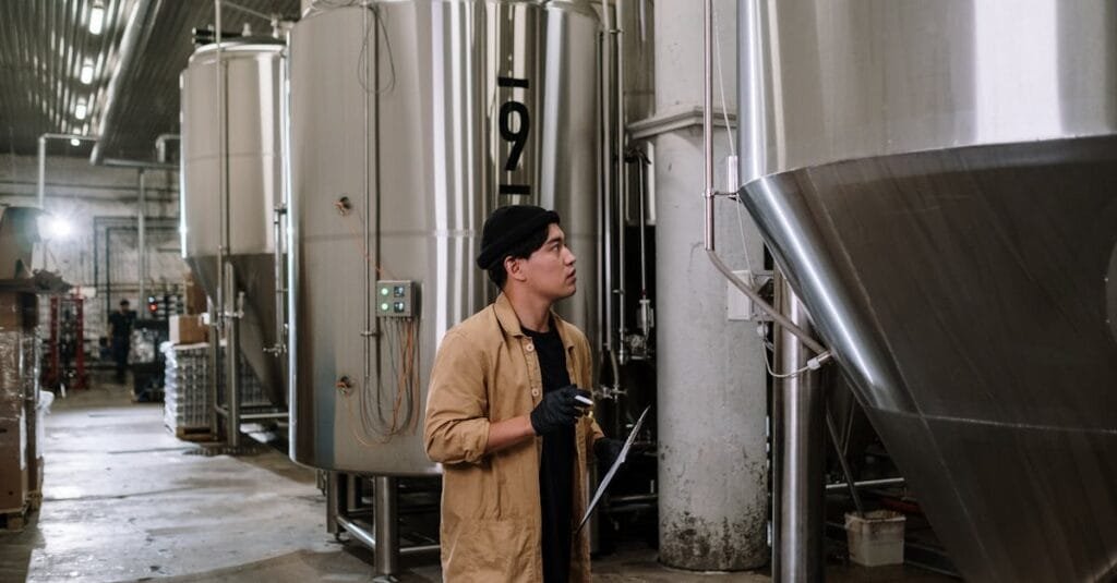 Asian man inspecting stainless steel tanks in a brewery, showcasing modern industrial processes.
