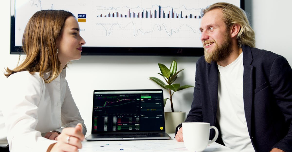 A business meeting with two professionals discussing data on laptops with charts displayed.
