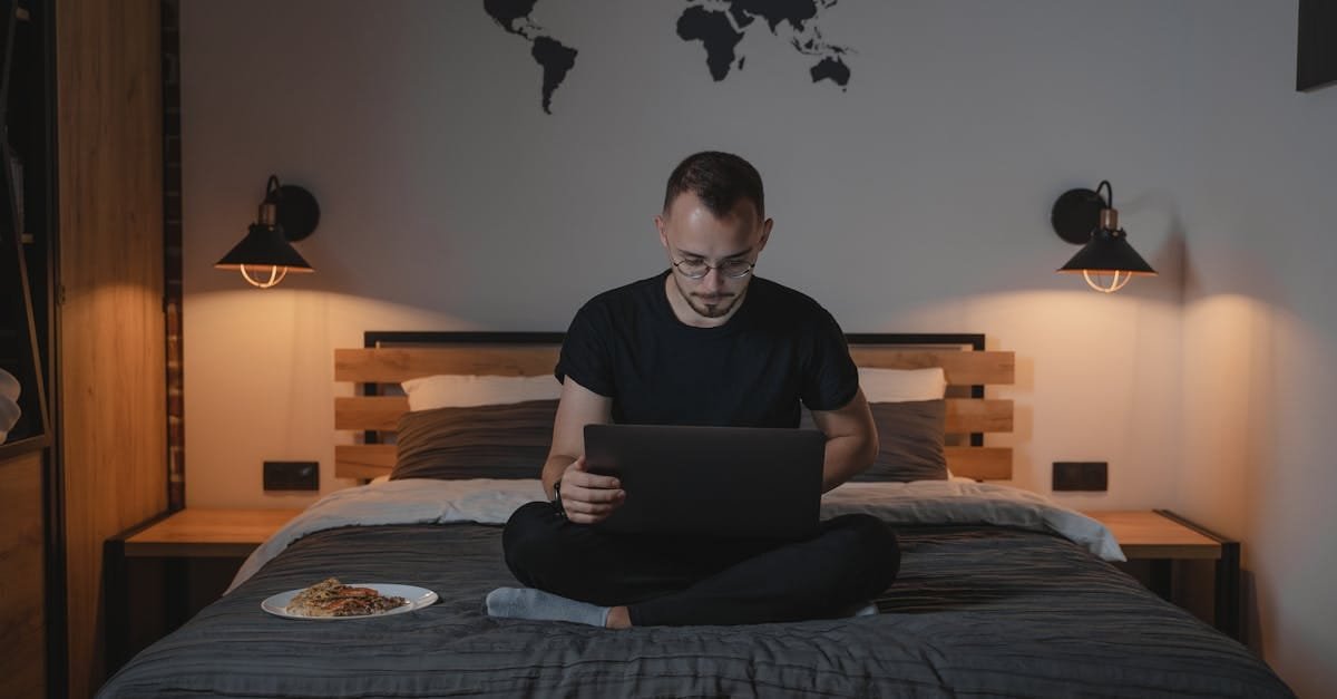 Man working on a laptop in a cozy bedroom setting, showcasing remote work lifestyle.