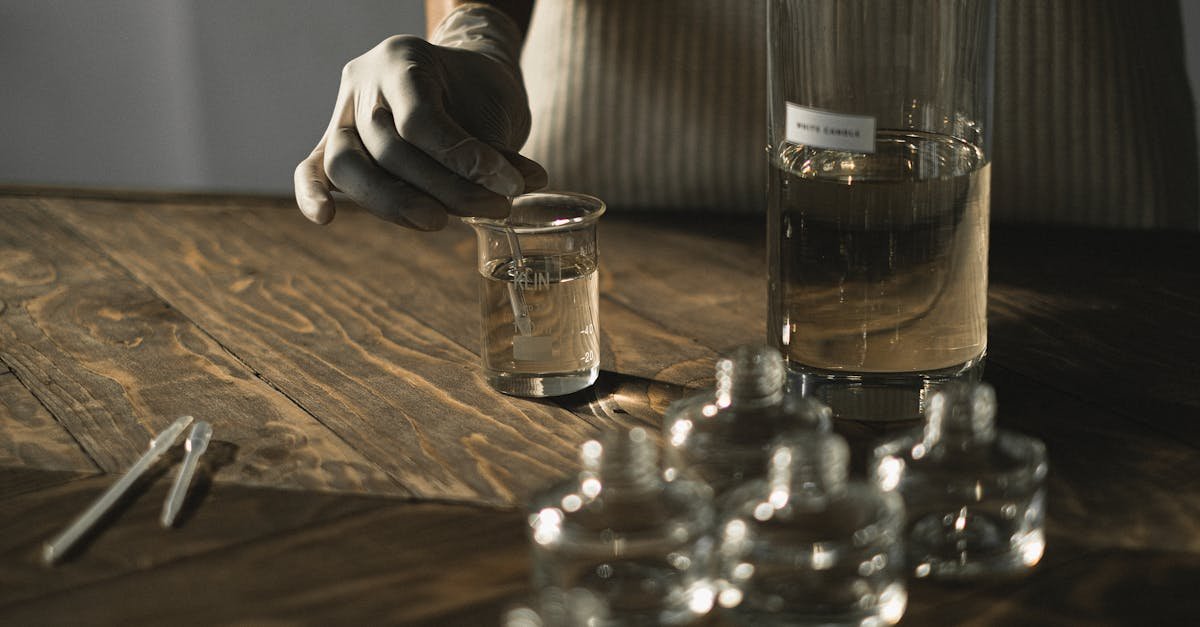 Crop anonymous female in latex gloves making fluid for perfume among glass jars on wooden table