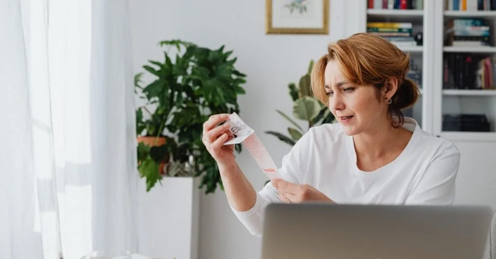 A woman in a white shirt sits indoors, examining a lengthy receipt with a concerned expression.