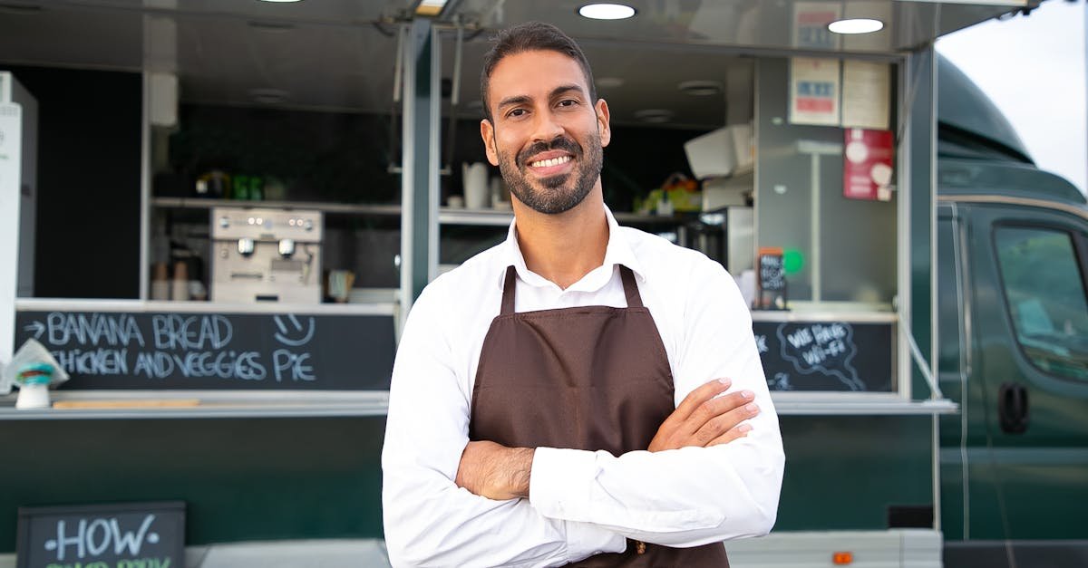 Smiling young ethnic male waiter in apron standing with arms folded near modern food truck and looking at camera contentedly