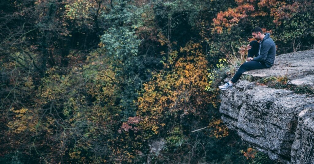A man sits pensively on a cliff edge surrounded by autumn foliage.