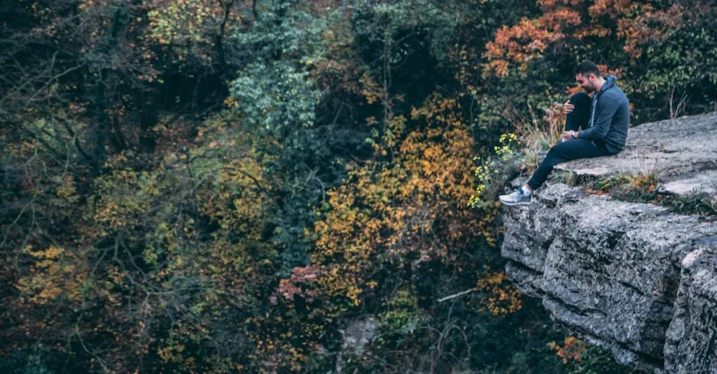 A man sits pensively on a cliff edge surrounded by autumn foliage.