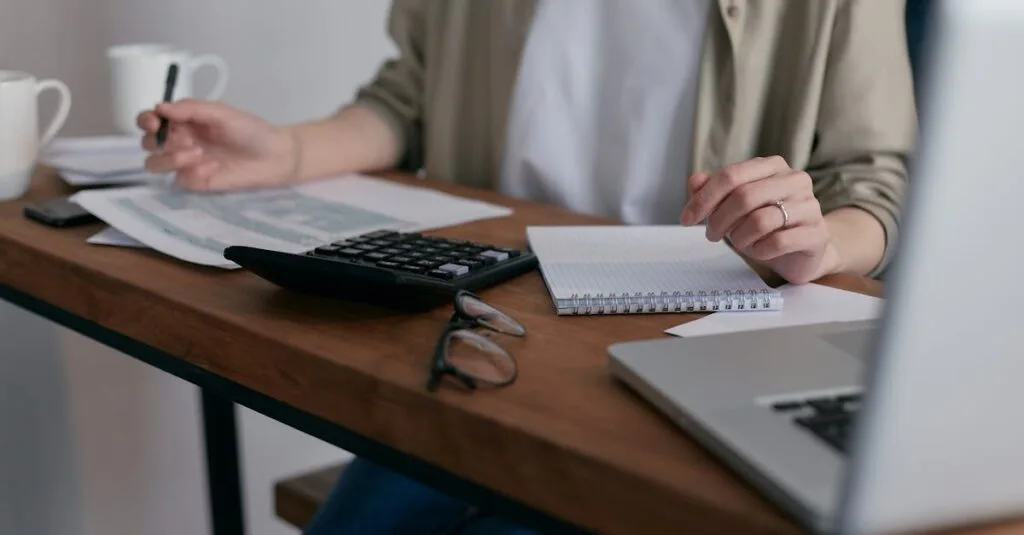 A woman manages finances at home, using a laptop and calculator on a wooden desk.
