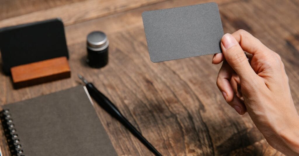 A close-up of a hand holding a blank business card above a wooden desk with office accessories.