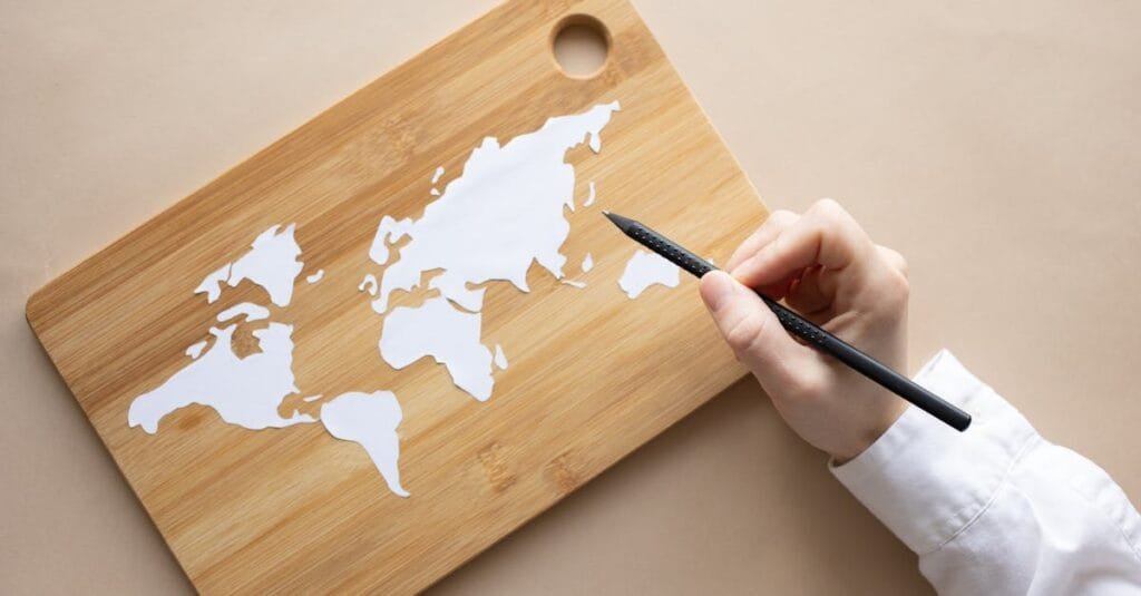 Hand crafting a world map on a bamboo cutting board using a pencil on beige background.
