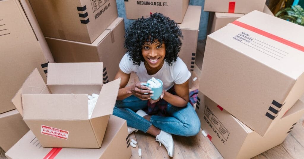 A woman sitting among moving boxes with a mug, surrounded by packing materials.