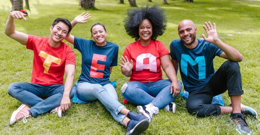 Multiracial team sitting on grass in park, wearing 'TEAM' shirts, showing unity.