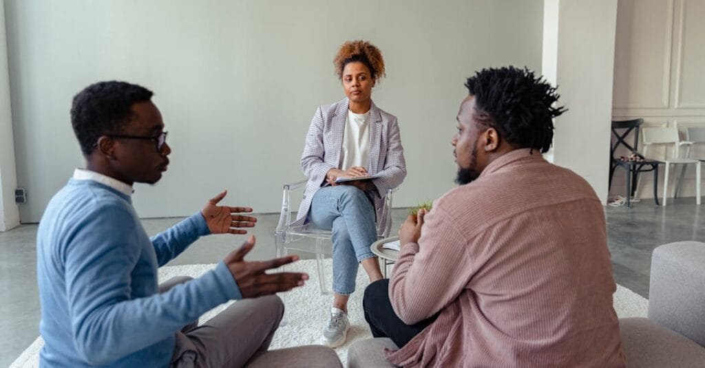 A therapist mediates a session with two men on a couch discussing mental health.