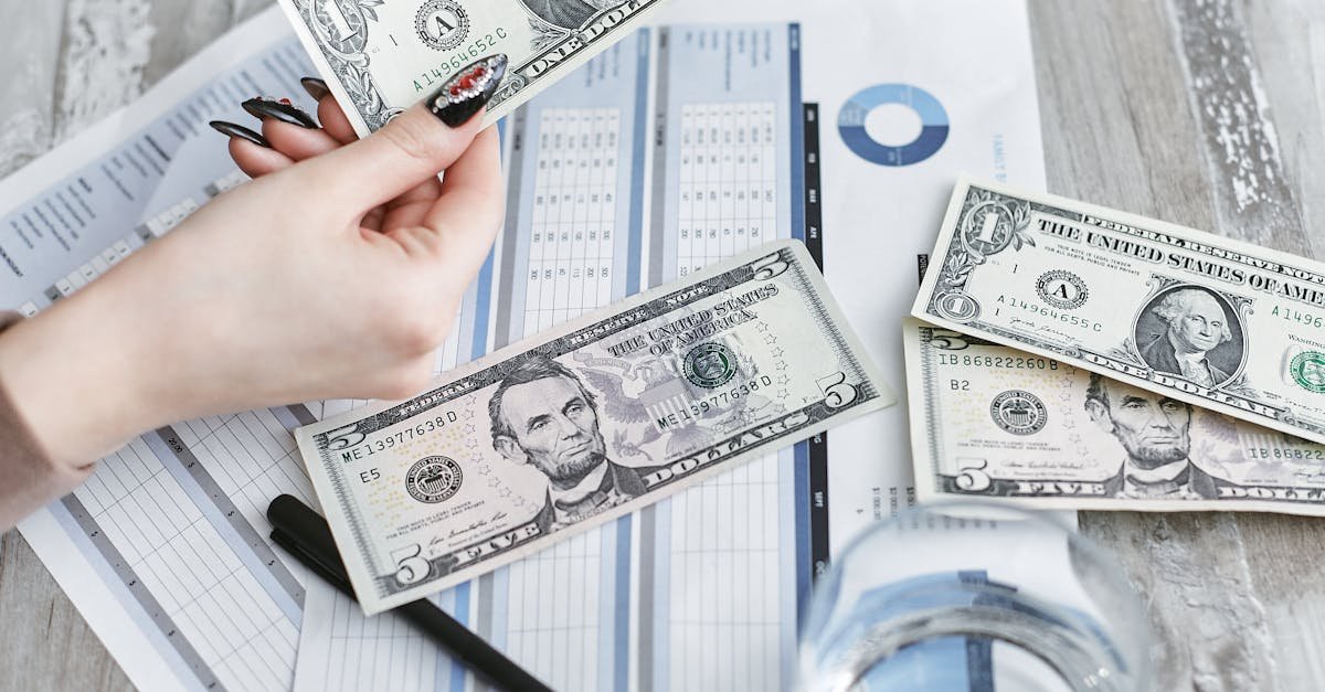 Close-up of a hand holding a dollar bill over financial documents and charts on a wooden table.