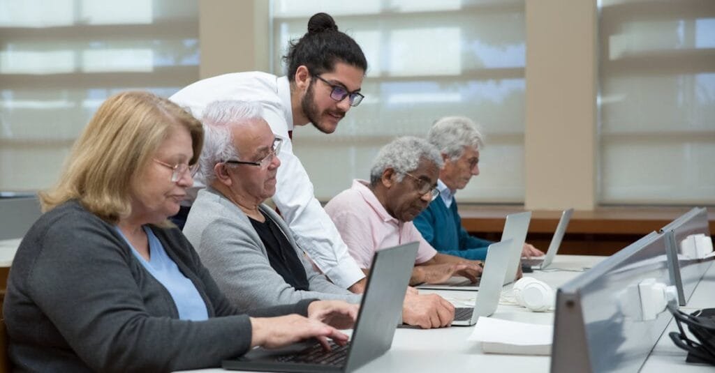 Senior adults use laptops in a classroom setting, guided by a young instructor.