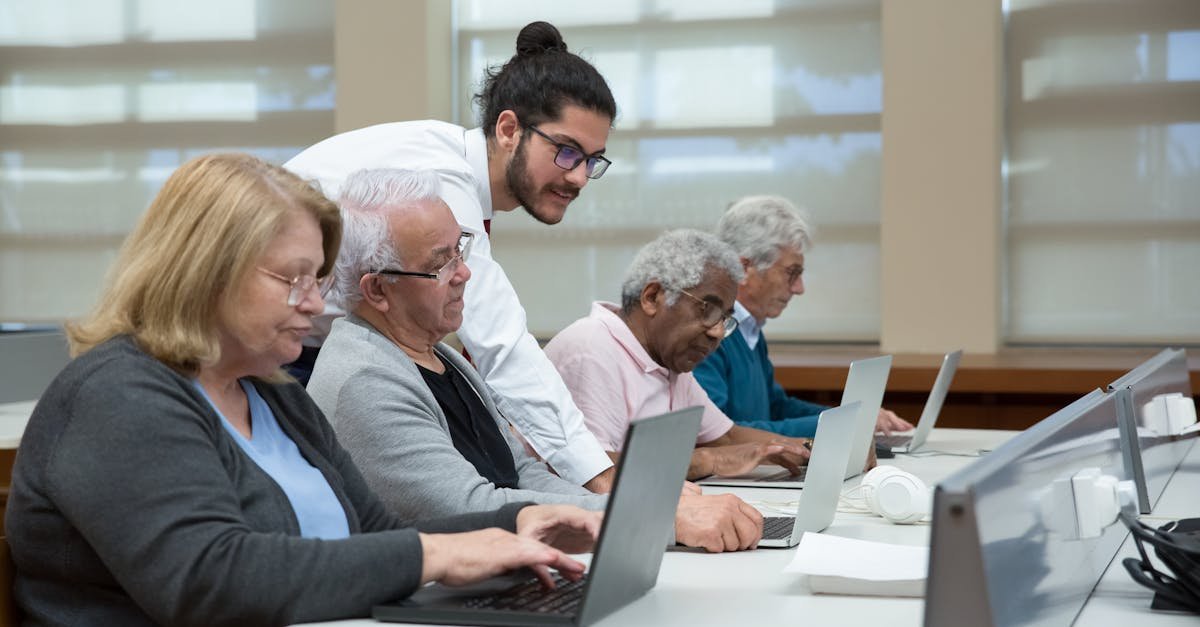 Senior adults use laptops in a classroom setting, guided by a young instructor.