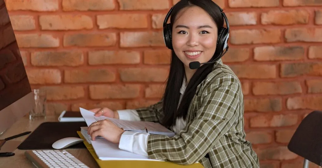 Asian woman working as a call center agent, smiling at desk with documents and headset.