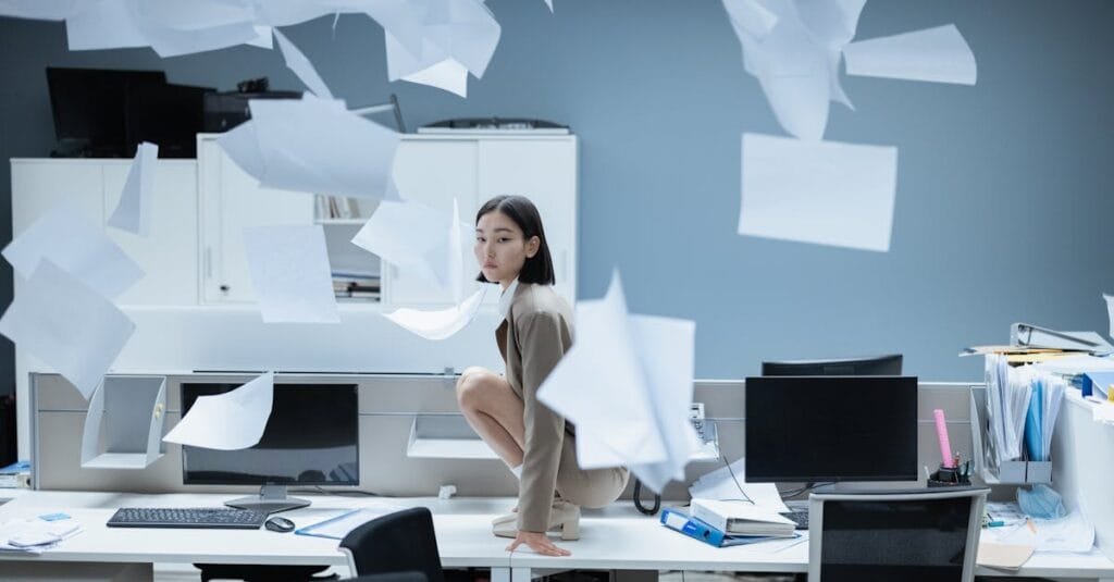 Woman perched on a desk in a chaotic office with papers flying, representing workplace stress.