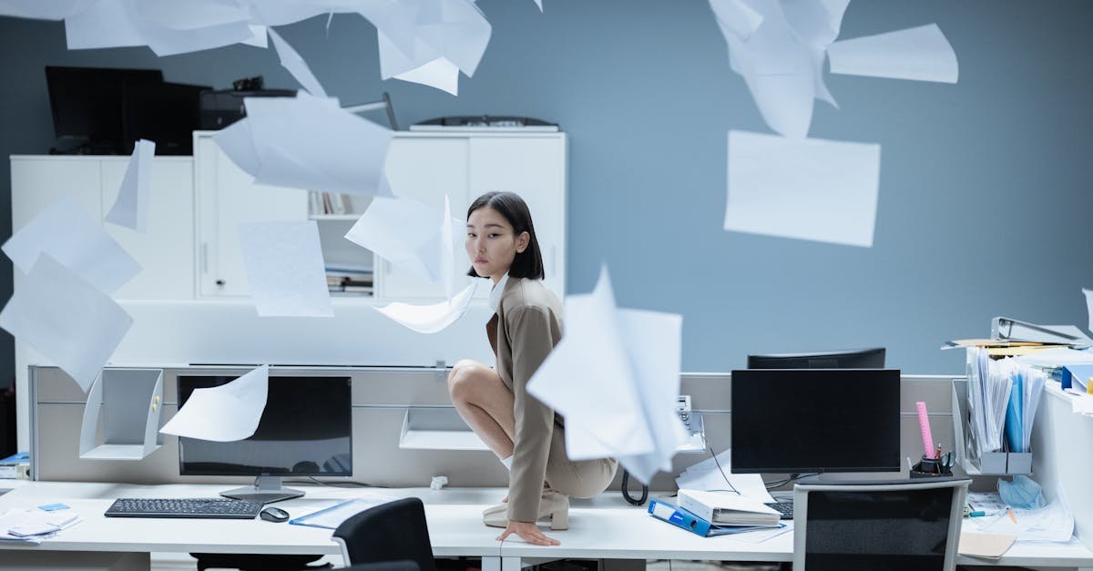 Woman perched on a desk in a chaotic office with papers flying, representing workplace stress.