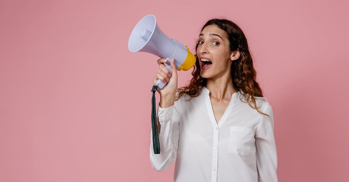 Woman in white shirt holding megaphone against a pink background, confidently speaking.