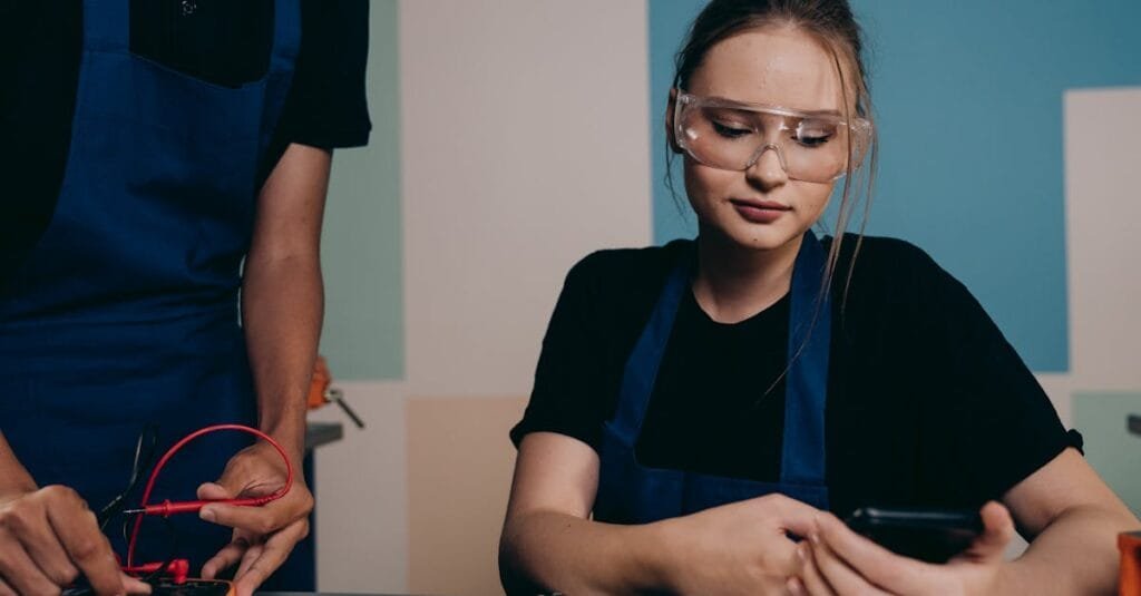 Two young engineers working with electronics, using smartphones and safety glasses for protection.