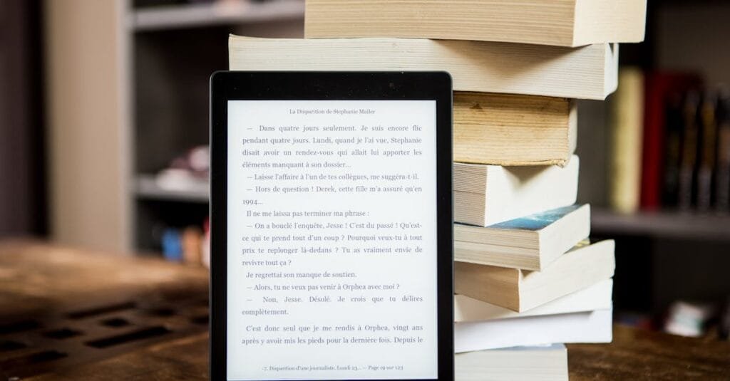 E-reader with text displayed next to a stack of physical books on a wooden table.