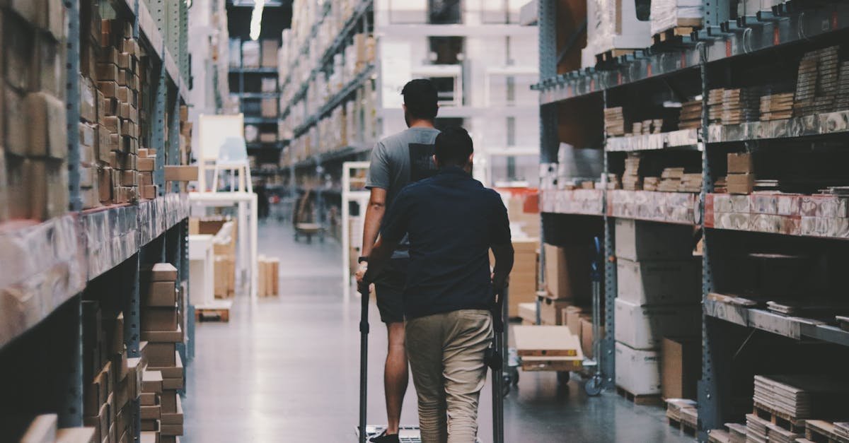 Two men maneuver a trolley in a large warehouse filled with boxes and shelves.