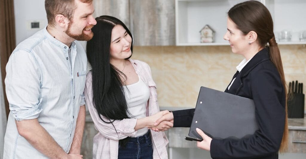 A real estate agent greets a couple with a handshake in a modern home interior.