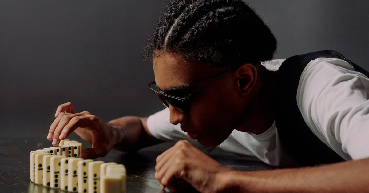 A man wearing sunglasses carefully lines up dominoes on a reflective surface, showing concentration and focus.